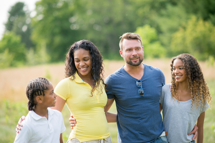 A happy family of four stands together in a sunny field, smiling and enjoying their time outdoors