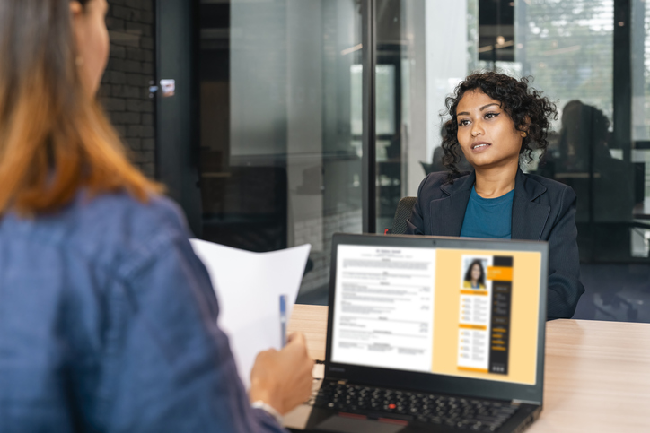 Two women seated at a desk during a professional meeting, with one reviewing documents on a laptop while the other listens.