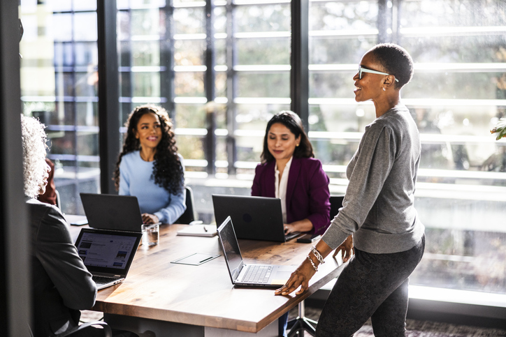 A group of colleagues gathered around a conference table during a collaborative meeting, with laptops open and one person standing and speaking.