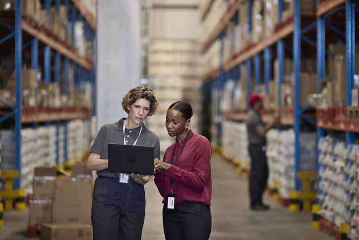 Two workers standing in a warehouse aisle reviewing information on a laptop, with storage shelves in the background.