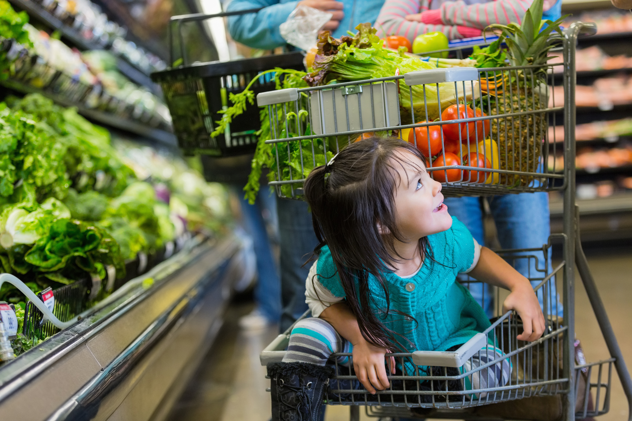 Elementary age Hispanic little girl is smiling while riding in bottom basket of shopping cart in supermarket.