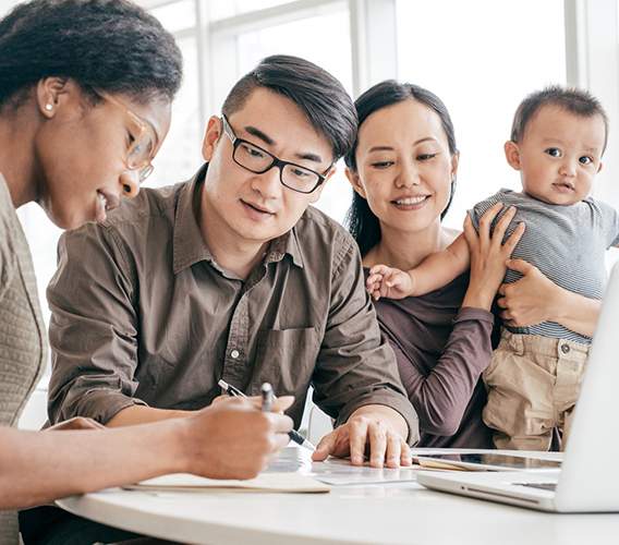 A family consisting of a man, woman, and baby sits at a table with a woman who appears to be assisting them, as they review documents and use a laptop in a bright, modern setting